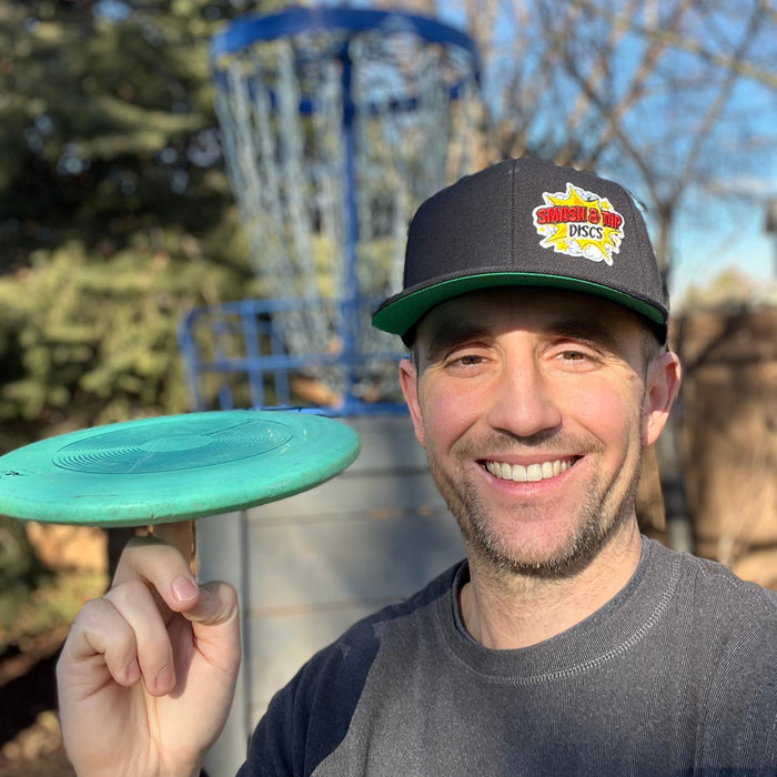 A smiling man outdoors wearing a dark gray and green cap with a 'Smash & Tap Discs' logo, holding a mint green disc golf disc on his finger. A disc golf basket is visible in the blurred background.